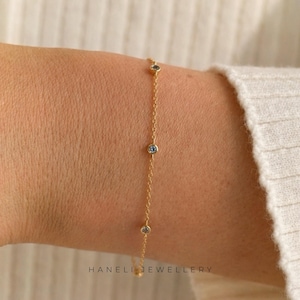 May include: A fine gold bracelet with three small blue gemstones set along a delicate chain. The bracelet is displayed on a wrist, with the brand name "HANELI JEWELLERY" printed at the bottom.