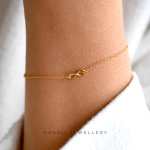 May include: A gold-coloured bracelet with an infinity symbol. The bracelet is attached to a fine chain. The brand name "HANELI JEWELLERY" is printed at the bottom of the image. The bracelet is worn on a person's wrist.
