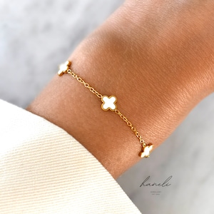 May include: Gold-coloured bracelet with four white clover charms. The fine chain links the charms, creating a simple yet elegant design. The bracelet is shown on a wrist, showcasing the jewellery's details and style.