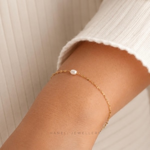 May include: A fine gold bracelet featuring a single, small, round pearl. The bracelet is worn on a wrist. The text "HANELI JEWELLERY" is at the bottom. The chain of the bracelet has a textured pattern.