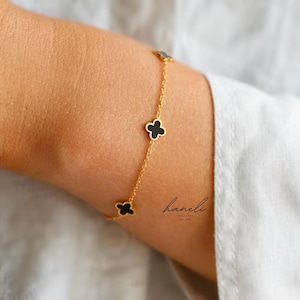 May include: A gold chain bracelet with four black clover-shaped charms. The bracelet is a delicate piece of jewellery. The brand name "haneli" is visible in script. The bracelet is worn on a wrist.