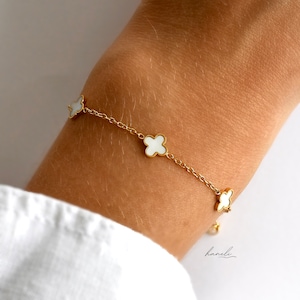 May include: A gold chain bracelet with white clover charms. The bracelet is on a person's wrist.