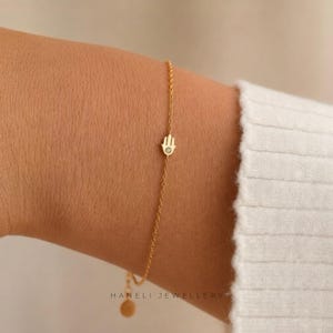 May include: A delicate gold bracelet with a Hamsa hand charm and a small gemstone. The bracelet is worn on a wrist, highlighting its fine chain and minimalist design. The text "HANELI JEWELLERY" is visible.