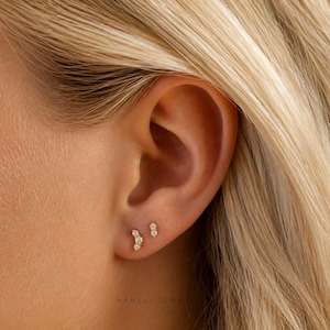 May include: Two gold stud earrings worn in the earlobe. One earring features a curved design with three small round clear stones, while the other is a single small round clear stone stud. The text Haneli Jewellers is visible at the bottom.