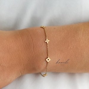 May include: A delicate gold bracelet with four-leaf clover and cross-shaped charms. The bracelet is worn on a wrist, with the brand name "haneli" in cursive script visible.