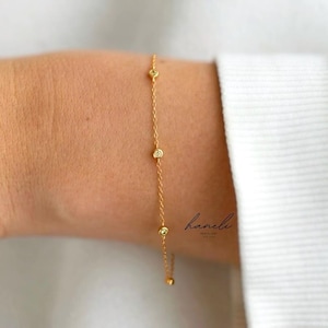 May include: A delicate gold bracelet with four small, round, sparkling accents. The bracelet is displayed on a person's arm, with a white shirt in the background. The bracelet is a simple, elegant piece of jewellery.