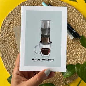 Op de afbeelding: Een wenskaart met een aquarel illustratie van een koffiezetapparaat boven een glazen mok gevuld met koffie. De kaart heeft de tekst "Happy brewday!" op een lichtblauwe achtergrond. De kaart wordt in een hand gehouden.