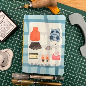 May include: A notebook with a blue and white checked cover, displaying running-themed illustrations such as a vest, shorts, and trainers. The text "RUNNING ERA UNLOCKED!" is visible. Other items include pens, a calendar, and a grey tool.