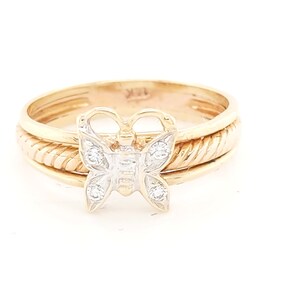 May include: A gold ring with a butterfly design. The butterfly has a silver-colored center with small, clear stones. The ring has a textured band and a smooth band. The ring is set against a white background.