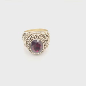 May include: A gold ring featuring a large, oval, red gemstone. The ring is inscribed with the words "VALLEY FORGE" around the central stone. The ring has detailed side engravings.