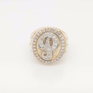 Op de afbeelding: Een gouden ring met een grote ovale zetting. De zetting is gevuld met kleine diamanten en heeft een grote letter "P" in het midden. De ring ligt op een witte achtergrond.