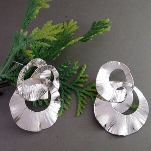 May include: A pair of silver-toned earrings with a textured, rippled design. Each earring features multiple interconnected circular shapes, creating a layered effect. The earrings are displayed on a gray surface with green foliage in the background.
