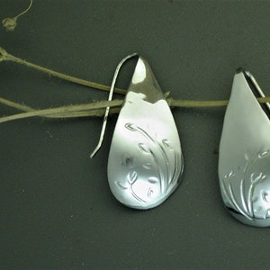 May include: A pair of silver teardrop earrings with a floral design. The earrings have a polished finish and a simple hook closure. The floral design is etched into the surface of the earrings. The earrings are displayed on a dark background.