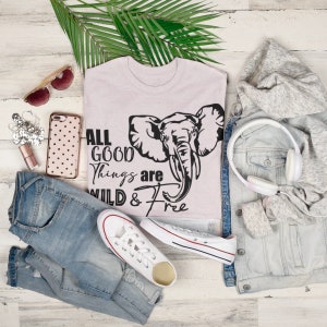 May include: A light pink t-shirt with a black elephant graphic and the text "All good things are wild & free". The shirt is laid out on a white wooden surface with a pair of jeans, a pair of white sneakers, a pair of sunglasses, a cell phone, a small bag of glitter, and a pair of headphones.