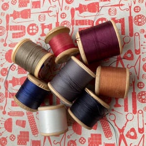 May include: A selection of wooden thread spools in a variety of colours, including brown, red, purple, grey, orange, blue, and white. The spools are arranged on a red and white patterned background, ideal for sewing and craft projects.