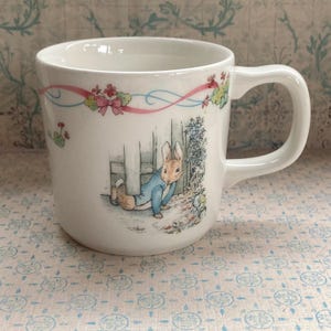 May include: White ceramic mug with a Peter Rabbit design. The illustration shows a rabbit in a blue jacket near a fence, with floral and ribbon details. The mug has a curved handle.