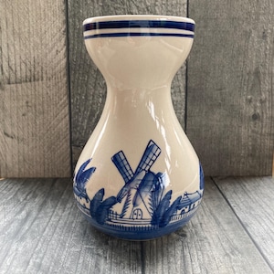 May include: A cream-coloured ceramic vase with a flared rim and a narrow neck. The body features a blue hand-painted design depicting a traditional windmill, foliage, and small buildings. Two horizontal blue bands encircle the top rim.