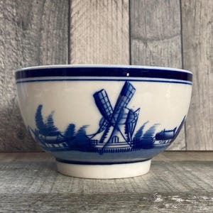 May include: White ceramic bowl featuring a blue windmill and landscape design. The bowl has a blue rim and a footed base. The windmill is the focal point, with detailed blue lines depicting the blades and structure.
