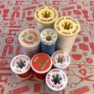 May include: A collection of nine sewing thread spools in various colours including white, blue, red, and orange. The spools feature plastic centres with printed text such as Molnlycke Sweden 100% Cotton, Drima, Star Dewhurst, and 110m 26nm labels.