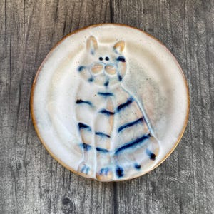 May include: A round ceramic plate with a cream-coloured glaze and a raised, embossed design of a cat. The cat figure features dark blue horizontal stripes across its body, a rounded face with two small ears, and two dark dots for eyes.