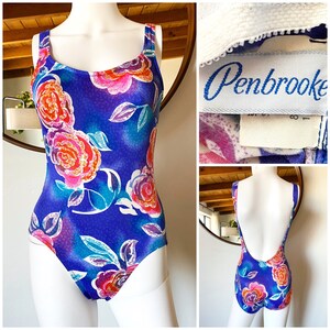 Op de afbeelding: Een blauwe badpak met een bloemenprint met roze, oranje en witte rozen. Het badpak heeft een ronde hals en een lage rug. Het label op het badpak luidt "Penbrooke".