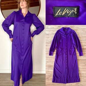 May include: A long, button-down purple dress with long sleeves, a collar, and two front pockets. The dress has a vintage style and is made of a shiny fabric. The tag reads "Le Voy's".