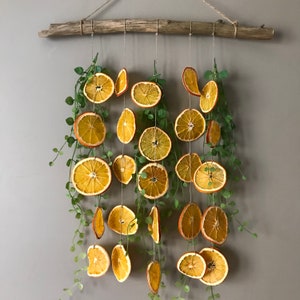 Puede incluir: Un tapiz de pared hecho de rodajas de naranja secas y vegetaci&oacute;n. Las rodajas de naranja est&aacute;n enhebradas en un cordel y colgadas de una rama de madera. La vegetaci&oacute;n est&aacute; intercalada entre las rodajas de naranja.