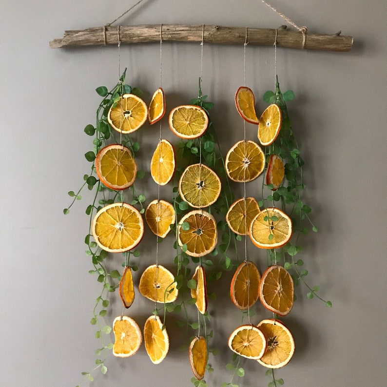 Puede incluir: Un tapiz de pared hecho de rodajas de naranja secas y vegetaci&oacute;n. Las rodajas de naranja est&aacute;n enhebradas en un cordel y colgadas de una rama de madera. La vegetaci&oacute;n est&aacute; intercalada entre las rodajas de naranja.