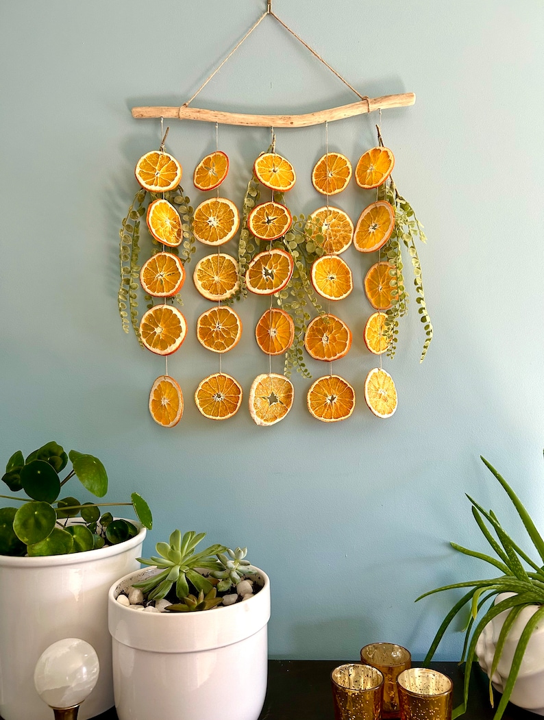 Puede incluir: Un tapiz de pared hecho de rodajas de naranja secas y vegetaci&oacute;n, colgado de una rama de madera flotante. Las rodajas de naranja est&aacute;n dispuestas en un patr&oacute;n en cascada, con la vegetaci&oacute;n intercalada. El tapiz de pared es una decoraci&oacute;n ligera y aireada que ser&iacute;a perfecta para un hogar con una est&eacute;tica r&uacute;stica o bohemia.