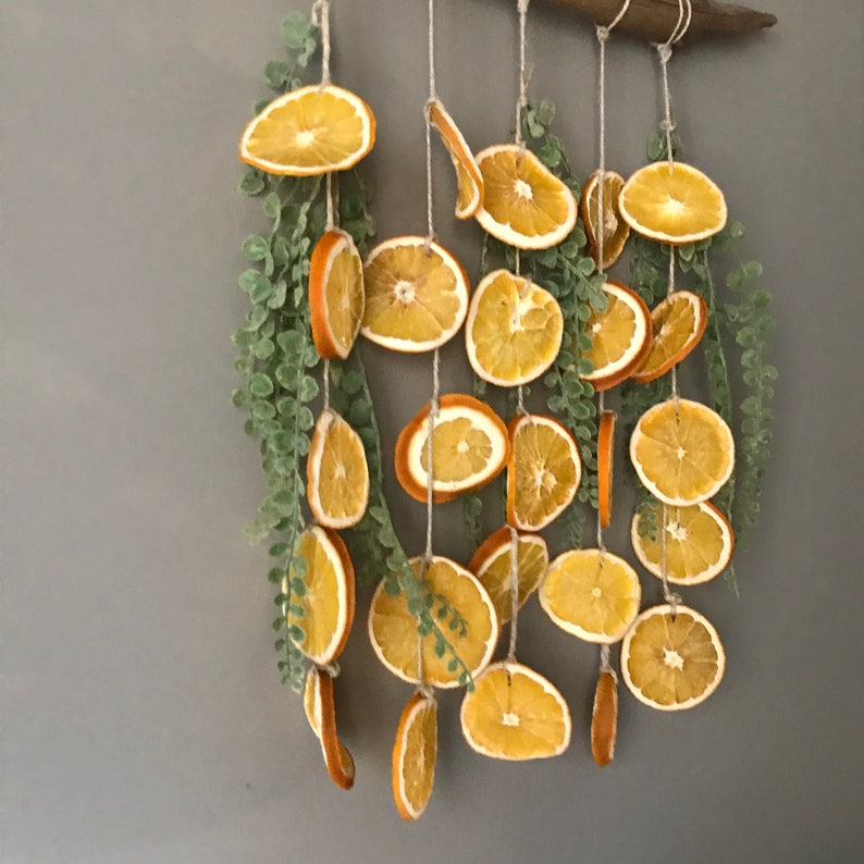 Puede incluir: Una guirnalda de rodajas de naranja secas con hojas verdes colgando de una varilla de madera. La guirnalda est&aacute; hecha de m&uacute;ltiples rodajas de naranja secas y hojas verdes, creando un aspecto r&uacute;stico y natural.