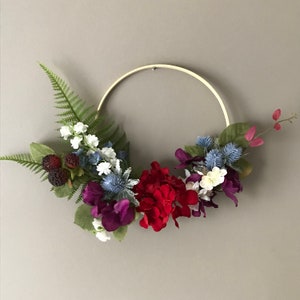 Puede incluir: Una corona de flores artificiales de color rojo, blanco y azul en un aro de madera. La corona presenta una variedad de flores, incluyendo hortensias, cardos y helechos.