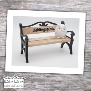 Puede incluir: Un pequeño banco de metal negro con un asiento de madera. El banco tiene un respaldo decorativo y las palabras "Lieblingsplatz" están grabadas en el asiento. Una figurita de búho blanca está sentada en el banco.