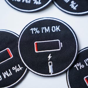 Puede incluir: Tres parches de hierro bordados en blanco y negro con el texto "1% I'M OK" y una imagen estilizada de una batería con una barra roja que indica una carga baja. La batería está conectada a un cable de carga.
