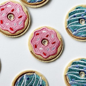 Może przedstawiać: Zestaw sześciu haftowanych naszywek do prasowania z motywami pączków. Pączki są w różnych kolorach, w tym różowym, niebieskim i białym. Każdy pączek ma biały lukier i posypki.