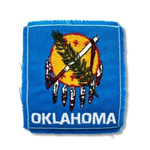 May include: A blue fabric patch with the state seal of Oklahoma. The seal features a yellow circle with a red and white handprint, a crossed spear and arrow, and a green sprig of willow. The word "OKLAHOMA" is embroidered in white below the seal.