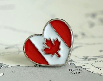 May include: A heart-shaped enamel pin featuring the Canadian flag design with a central red maple leaf on a white background, flanked by two red vertical stripes, all enclosed within a polished silver-toned metal border.