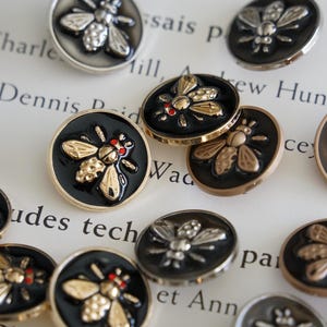 Peut inclure: Un ensemble de boutons décoratifs avec un motif d'abeille. Les boutons sont ronds, avec un fond en émail noir et des détails d'abeille dorés ou argentés. Certaines abeilles ont des accents rouges. Les boutons sont disposés sur une surface blanche.