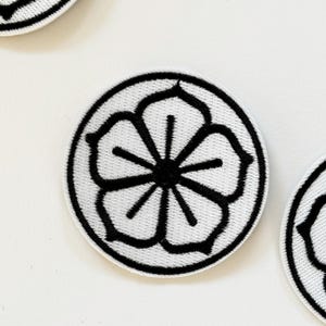 May include: White embroidered patch with a black flower design. The flower has five petals and a centre circle.