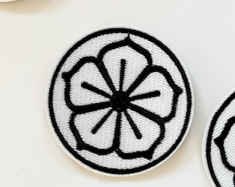 May include: White embroidered patch with a black flower design. The flower has five petals and a center circle.