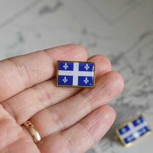 May include: A gold-colored pin with a blue and white flag of Quebec. The flag features a white cross with four fleur-de-lis on a blue background.