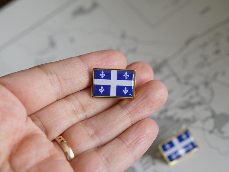 May include: A gold-colored pin with a blue and white flag of Quebec. The flag features a white cross with four fleur-de-lis on a blue background.