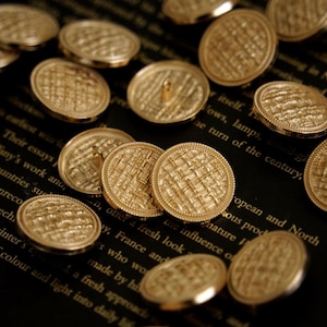 Peut inclure: Gros plan de plusieurs boutons dorés avec un motif de texture tissée. Les boutons ronds ont un bord légèrement surélevé et une boucle au dos pour la couture. Ils sont disposés sur une surface sombre avec du texte.