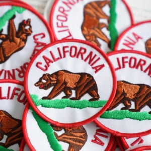 Könnte beinhalten: Eine Sammlung runder Aufnäher mit der kalifornischen Staatsflagge. Jeder Aufnäher hat einen weißen Hintergrund mit einem braunen Bären, grünem Gras und dem Wort "CALIFORNIA" in Rot. Die Aufnäher sind rot umrandet.