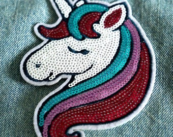 May include: A unicorn-shaped iron-on patch featuring white, teal, purple, and dark red sequins. The patch depicts a unicorn head in profile with a closed eye, a single horn, and a flowing mane, all outlined with a white fabric border.