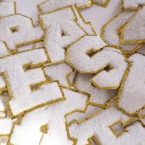 May include: Close-up of white felt letter patches with gold glitter trim. The letters are in a varsity style and are scattered across a white surface. The patches are likely for crafting or personalisation projects.