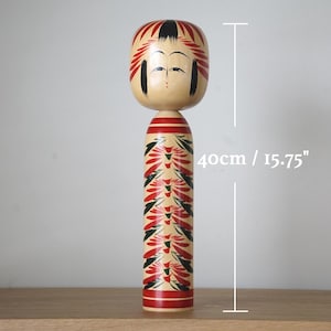 Könnte beinhalten: Eine traditionelle Kokeshi-Puppe, etwa 40 cm hoch, mit cremefarbenem Körper und bemaltem Gesicht. Der Kopf der Puppe hat rot-schwarze Details, und der Körper ist mit einem rot-schwarzen Blumenmuster verziert.
