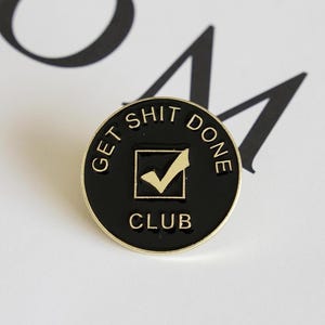 May include: A round, black enamel pin with gold trim. The pin features the text "GET SHIT DONE CLUB" around the edge. In the center is a gold checkmark inside a square. The pin is a motivational accessory.