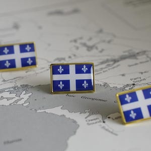 May include: Three gold pins with the flag of Quebec on a map of Canada. The flag is blue with a white cross and four fleur-de-lis.