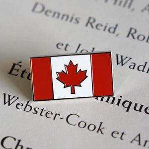 Peut inclure: Épinglette rectangulaire représentant le drapeau canadien. Le design présente des barres rouges verticales encadrant un panneau central blanc avec une feuille d'érable rouge. L'épinglette est posée sur un texte imprimé.