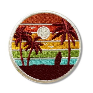 May include: A round embroidered patch depicting a sunset scene with palm trees, a surfboard, and a white moon. The background features a multi-coloured striped pattern in shades of orange, yellow, green, and blue.